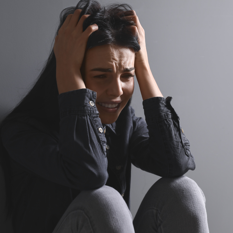 A distressed woman sitting against a gray wall, holding her head in her hands, reflecting feelings of uncertainty and worry about the year 2025.