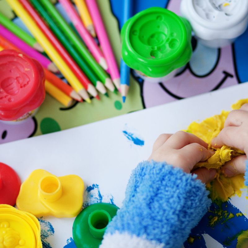 A child's hands molding yellow clay, surrounded by colorful pencils and paints, representing the playful and exploratory nature of creativity born from uncertainty.