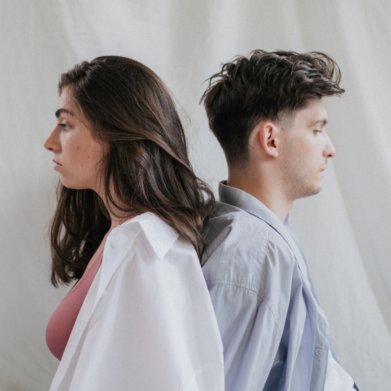A man and woman sit back-to-back, both looking away with closed eyes and solemn expressions, suggesting emotional disconnection and unresolved tension and unsure of what they feel.