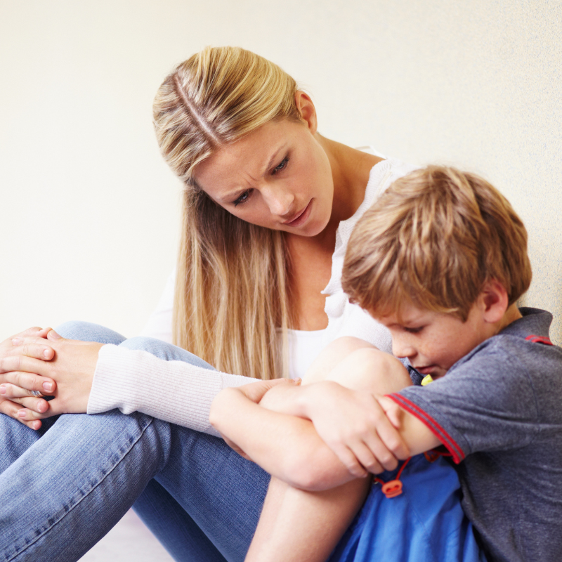 A young boy sitting with his knees pulled to his chest, showing visible emotion, while a concerned woman gently listens and offers support.