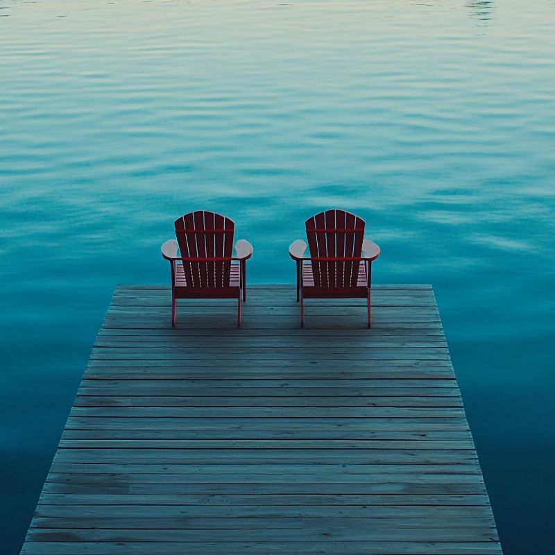 Two empty red chairs on a dock overlooking calm water, representing friendship, remembrance, and love that endure beyond loss.